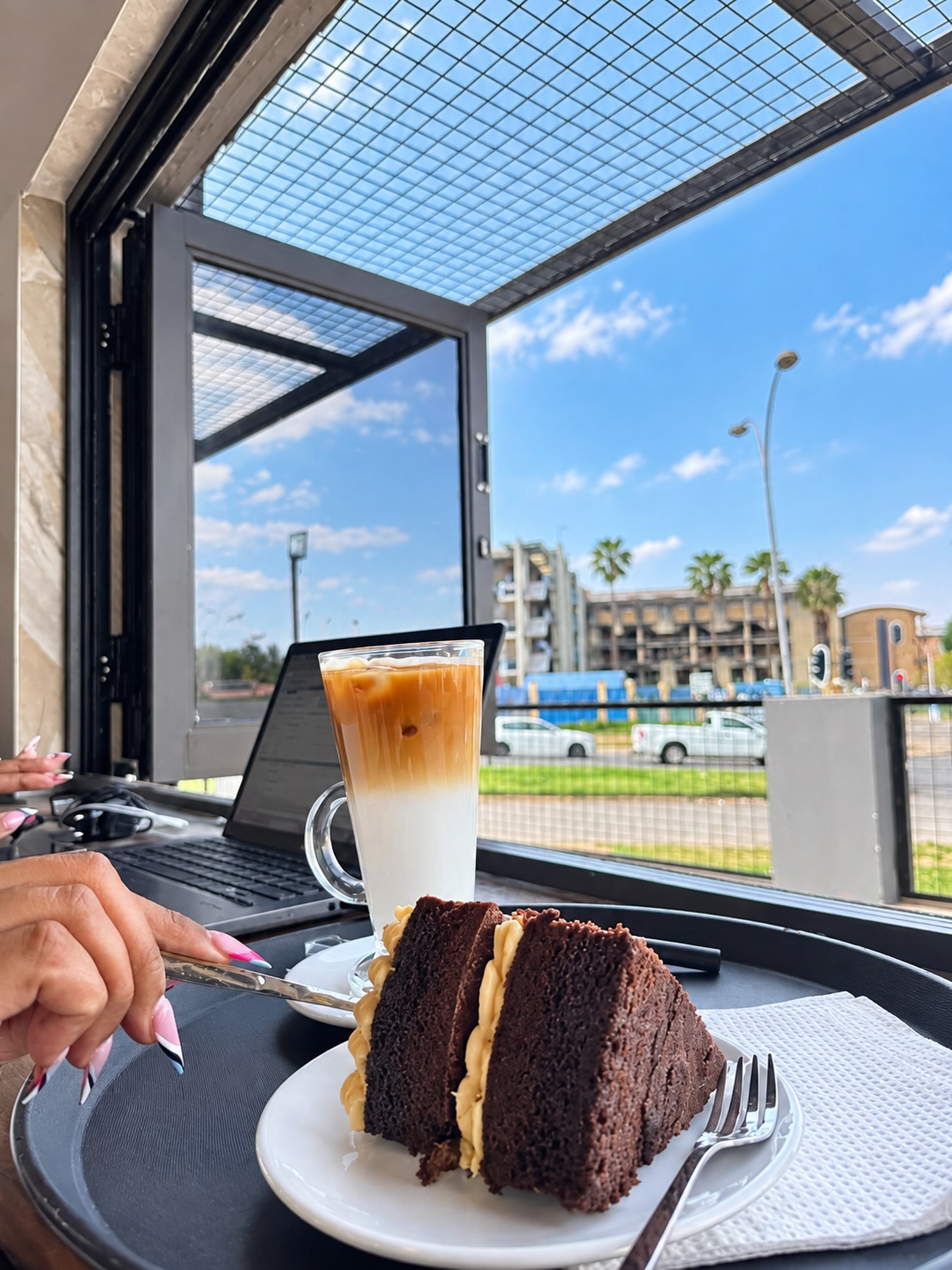 An iced latte and a slice of chocolate cake at the window bar, with a laptop open alongside